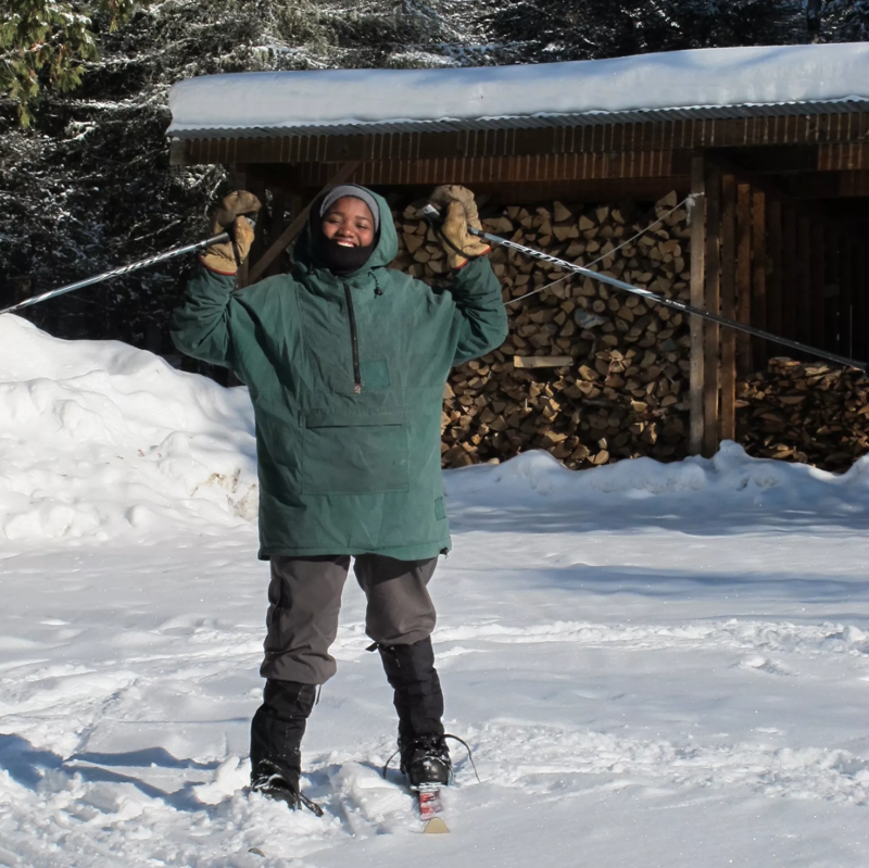 A person stands in the snow, wearing skis and holding ski poles up in the air with a big smile. They are dressed in a green hooded jacket, grey pants, and black boots. Behind them is a wooden structure filled with firewood, and snow-covered ground and trees surround the scene. The sun is shining, creating a bright and cheerful winter atmosphere.